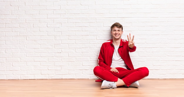 Giovane uomo biondo che sorride e che sembra amichevole, mostrando numero tre o terzo con la mano in avanti, contando alla rovescia seduta sul pavimento