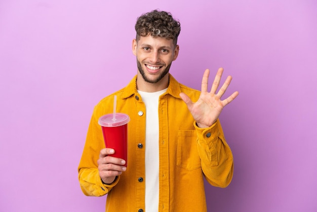Giovane uomo biondo che tiene soda isolata su sfondo viola contando cinque con le dita