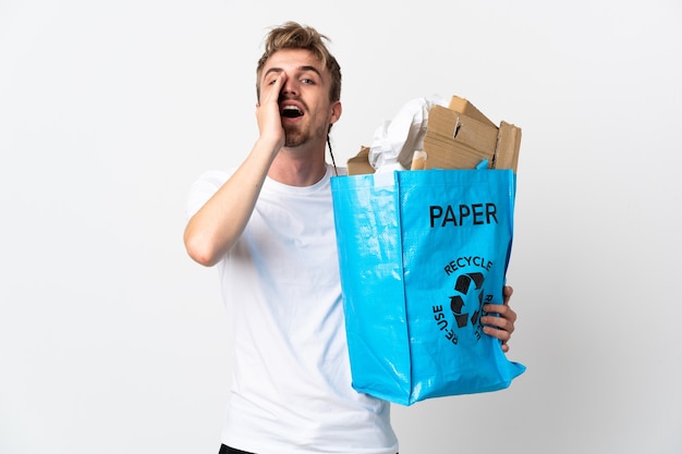 Giovane uomo biondo che tiene un sacchetto di riciclaggio pieno di carta da riciclare isolato su sfondo bianco gridando e annunciando qualcosa