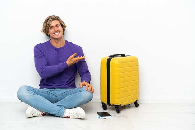 Giovane uomo biondo con la valigia che si siede sul pavimento che presenta un'idea mentre guarda sorridente verso
