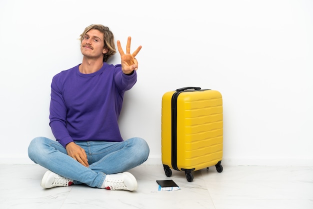 Giovane uomo biondo con la valigia che si siede sul pavimento felice e contando tre con le dita