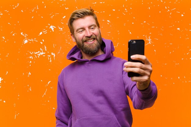 Giovane uomo biondo con uno smart phone che indossa una felpa con cappuccio viola contro il muro arancione danneggiato