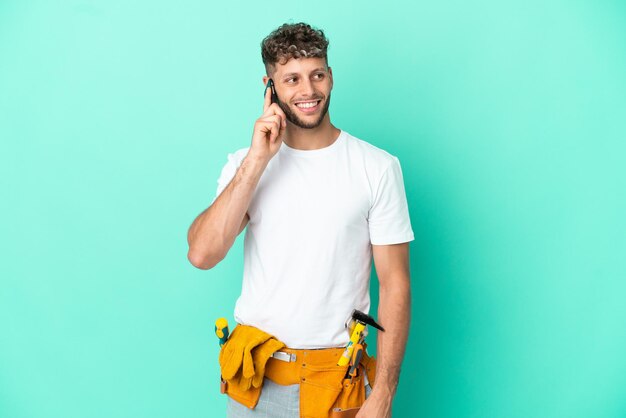 Giovane uomo biondo elettricista isolato su sfondo verde che tiene una conversazione con il telefono cellulare