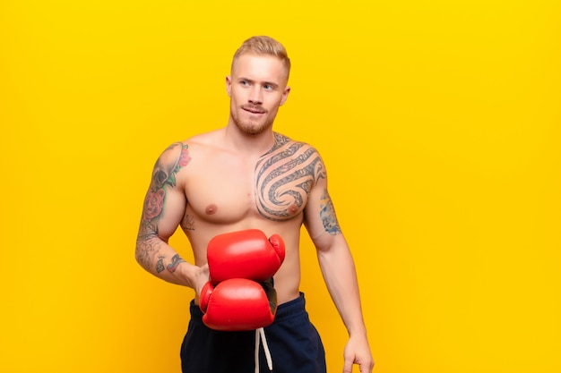 Giovane uomo biondo forte che dubita o pensa, morde le labbra e si sente insicuro e nervoso, cercando di copiare lo spazio sul lato con guantoni da boxe