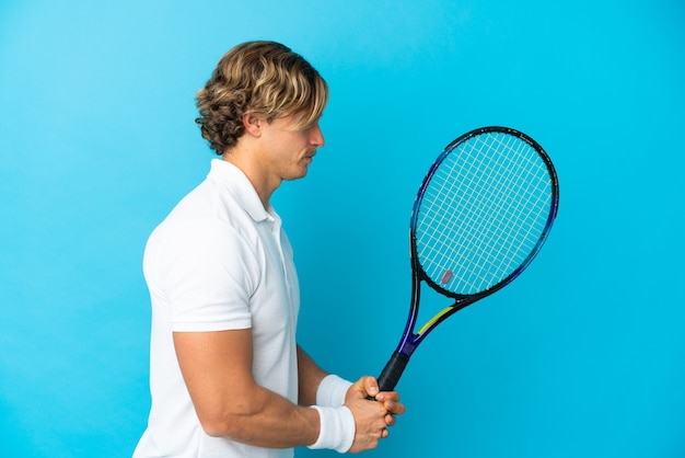 Giovane uomo biondo isolato sulla parete blu giocando a tennis