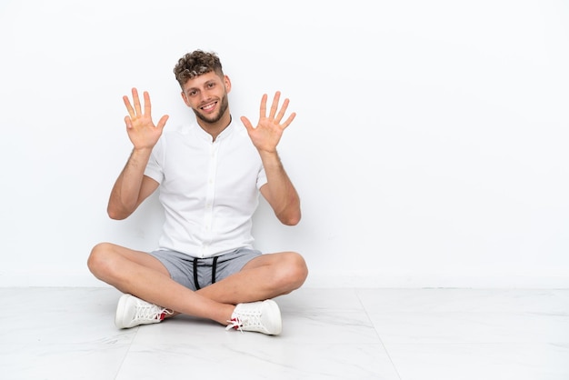 Giovane uomo biondo seduto sul pavimento isolato su sfondo bianco contando nove con le dita