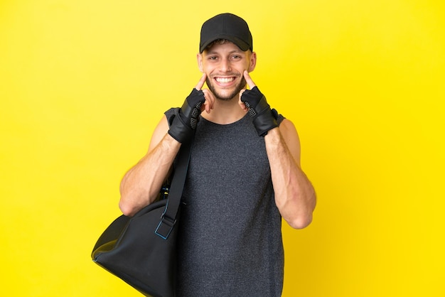 Giovane uomo biondo sportivo con borsa sportiva isolata su sfondo giallo sorridente con un'espressione felice e piacevole