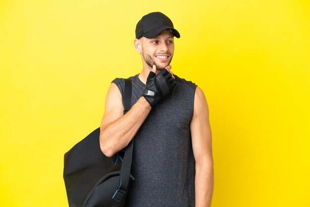 Giovane uomo biondo sportivo con borsa sportiva isolato su sfondo giallo che guarda al lato