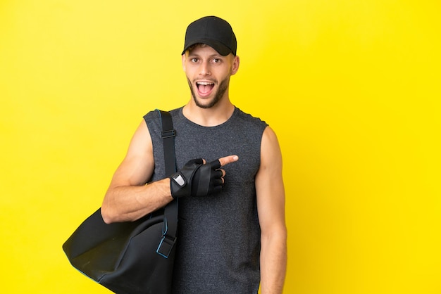 Giovane uomo biondo sportivo con borsa sportiva isolato su sfondo giallo sorpreso e rivolto verso il lato