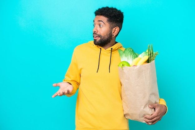 Giovane uomo brasiliano che prende un sacchetto di cibo da asporto isolato su sfondo blu con espressione sorpresa mentre guarda di lato