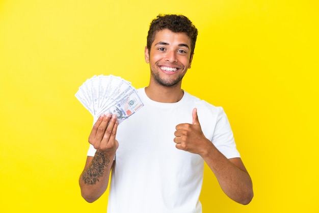 Giovane uomo brasiliano che prende un sacco di soldi isolato su sfondo giallo dando un gesto di pollice in alto