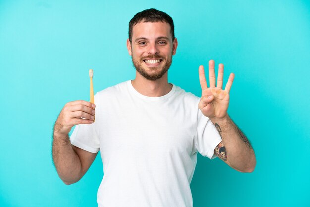 Giovane uomo brasiliano che si lava i denti isolati su sfondo blu felice e conta quattro con le dita