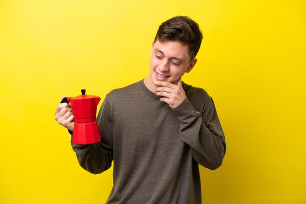 Giovane uomo brasiliano che tiene caffettiera isolata su sfondo giallo che guarda al lato e sorridente