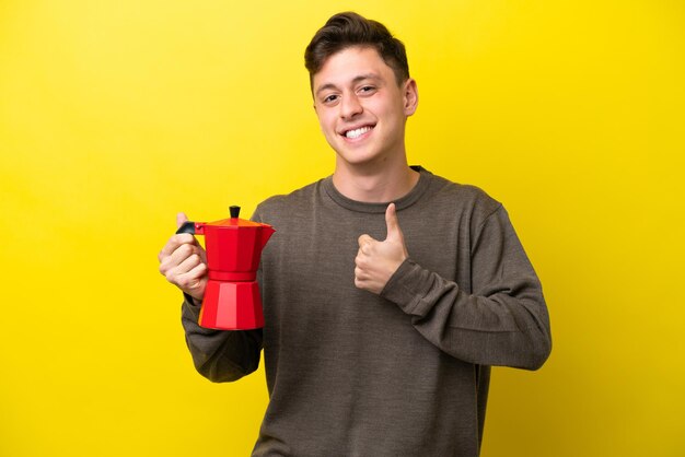 Giovane uomo brasiliano che tiene caffettiera isolata su sfondo giallo dando un pollice in alto gesto