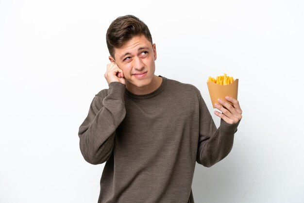 Giovane uomo brasiliano che tiene patatine fritte isolate su sfondo bianco frustrato e copre le orecchie