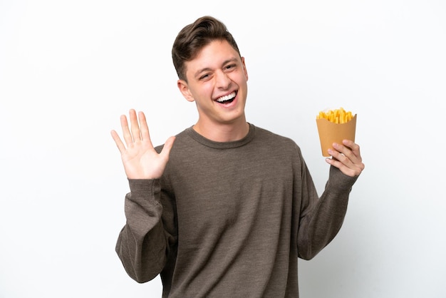 Giovane uomo brasiliano che tiene patatine fritte isolate su sfondo bianco salutando con la mano con espressione felice