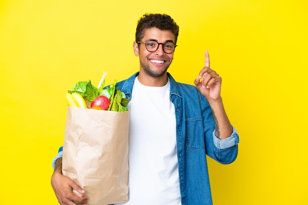 Giovane uomo brasiliano che tiene una borsa della spesa isolata su sfondo giallo che indica una grande idea