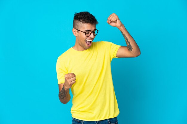 Giovane uomo brasiliano isolato su uno spazio blu che celebra una vittoria