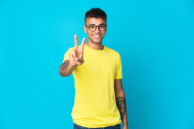 Giovane uomo brasiliano isolato sulla parete blu che sorride e che mostra il segno di vittoria