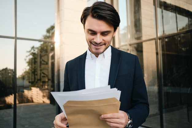 Giovane uomo bruna barbuto sorridente attraente in camicia bianca e giacca classica in piedi all'aperto mentre legge felicemente documenti con edificio in vetro sullo sfondo