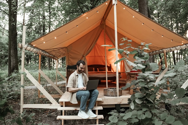 Giovane uomo bruna sorridente seduto sul glamping utilizzando il computer portatile, digitando, messaggistica nei social media. Stile di vita da campeggio. Viaggi a basso budget. Concetto di lavoro a distanza. Moderna tecnologia di comunicazione Internet.