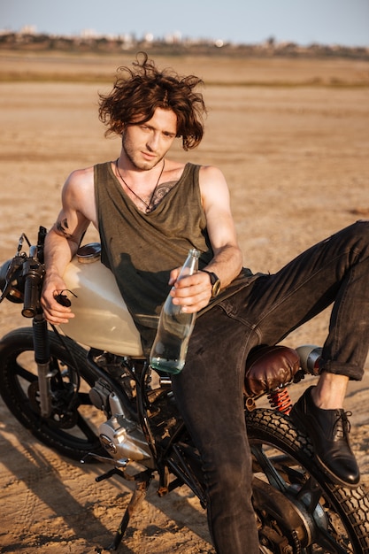 Giovane uomo brutale sdraiato sulla sua moto nel deserto e con in mano una bottiglia d'acqua di vetro