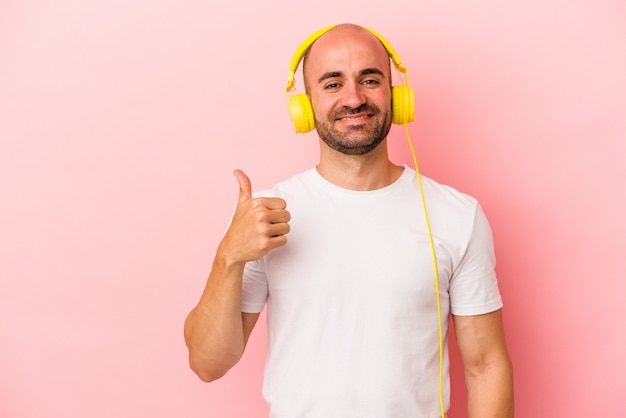 Giovane uomo calvo caucasico che ascolta musica isolato su sfondo rosa sorridente e alzando il pollice