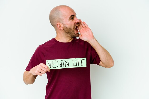 Giovane uomo calvo caucasico che tiene un cartello di vita vegano isolato su sfondo viola gridando e tenendo il palmo vicino alla bocca aperta.
