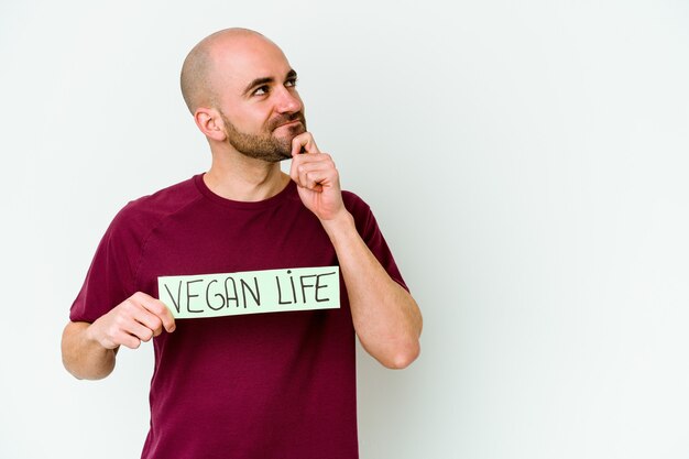 Giovane uomo calvo caucasico che tiene un cartello di vita vegano isolato sul muro viola che guarda lateralmente con espressione dubbiosa e scettica