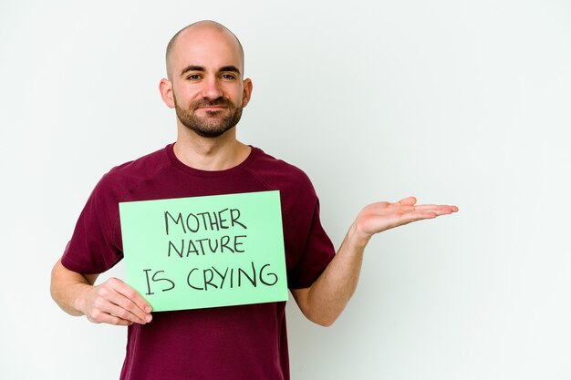 Giovane uomo calvo caucasico che tiene un pianto di madre natura isolato sulla parete bianca che mostra uno spazio della copia su una palma e che tiene un'altra mano sulla vita.