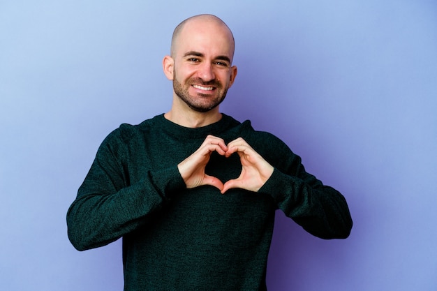 Giovane uomo calvo caucasico isolato su viola sorridente e mostrando una forma di cuore con le mani.