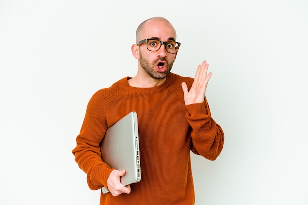 Giovane uomo calvo che tiene un computer portatile isolato sul muro bianco sorpreso e scioccato.