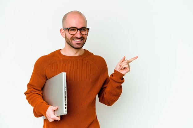 Giovane uomo calvo che tiene un computer portatile isolato sul muro bianco sorridendo e indicando da parte, mostrando qualcosa in uno spazio vuoto.