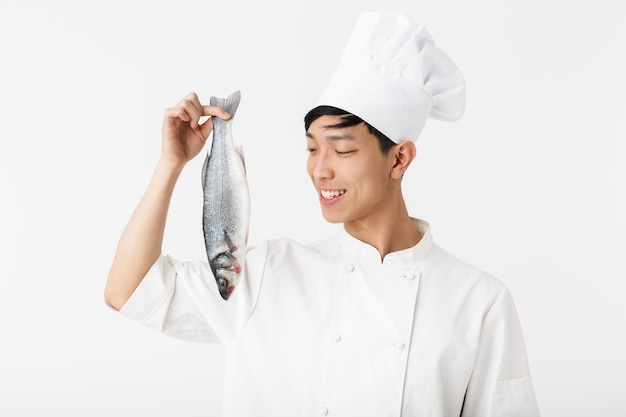 giovane uomo capo positivo in uniforme da cuoco bianca e cappello sorridente mentre si tiene il pesce fresco crudo isolato sul muro bianco