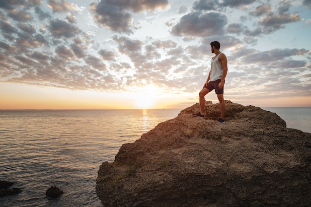 Giovane uomo casual in piedi sulla roccia di montagna in riva al mare e guardando il tramonto