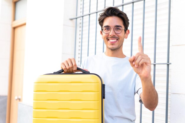 Giovane uomo caucasico all'aperto in vacanza con la valigia da viaggio e contando uno