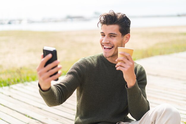 Giovane uomo caucasico all'aperto utilizzando il telefono cellulare e tenendo un caffè con un'espressione felice