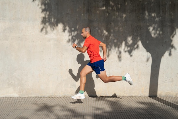 Giovane uomo caucasico atletico che corre accanto al muro di cemento L'ombra è proiettata sul muro Indossando maglietta rossa e pantaloncini blu Spagna Alicante