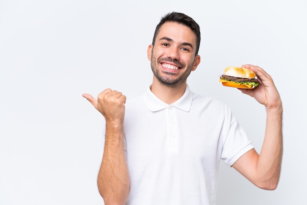 Giovane uomo caucasico bello che tiene un hamburger sopra fondo isolato che indica il lato per presentare un prodotto