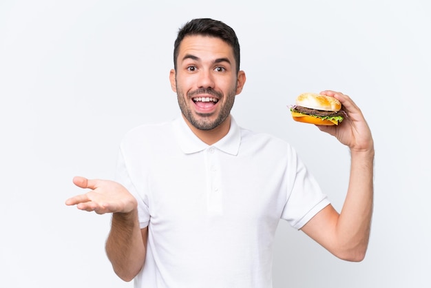Giovane uomo caucasico bello che tiene un hamburger su sfondo isolato con espressione facciale scioccata