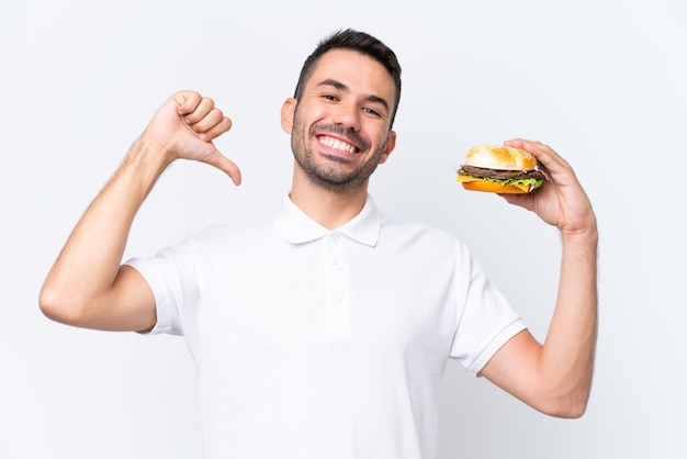 Giovane uomo caucasico bello che tiene un hamburger su sfondo isolato orgoglioso e soddisfatto di sé