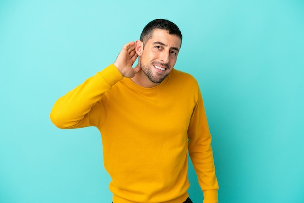 Giovane uomo caucasico bello isolato su sfondo blu ascoltando qualcosa mettendo la mano sull'orecchio