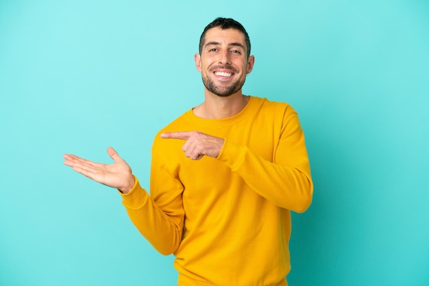 Giovane uomo caucasico bello isolato su sfondo blu che tiene copyspace immaginario sul palmo per inserire un annuncio