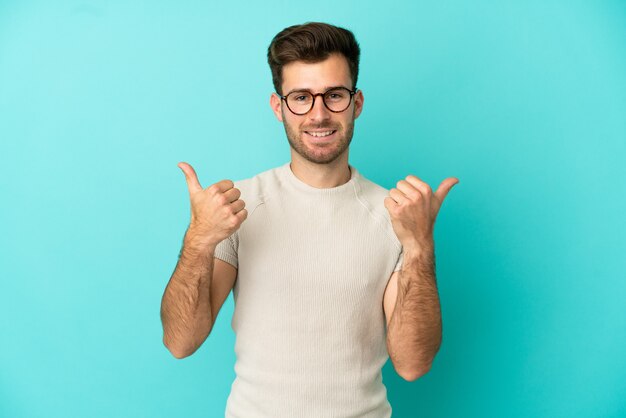 Giovane uomo caucasico bello isolato su sfondo blu con il pollice in alto gesto e sorridente