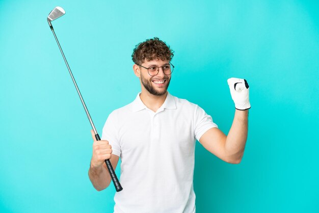 Giovane uomo caucasico bello isolato su sfondo blu, giocando a golf e celebrando una vittoria