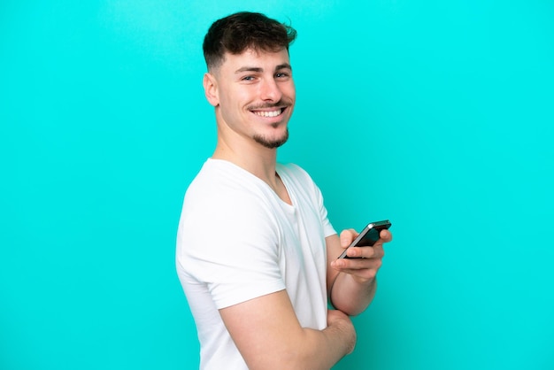 Giovane uomo caucasico bello isolato su sfondo blu in possesso di un telefono cellulare e con le braccia incrociate