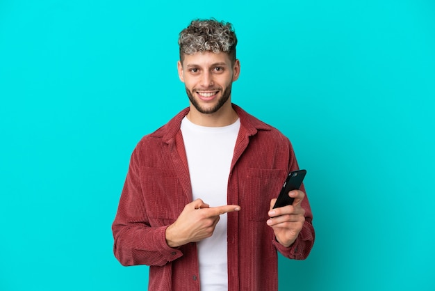 Giovane uomo caucasico bello isolato su sfondo blu usando il telefono cellulare e puntandolo