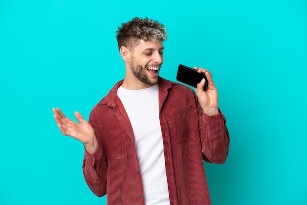 Giovane uomo caucasico bello isolato su sfondo blu utilizzando il telefono cellulare e cantando
