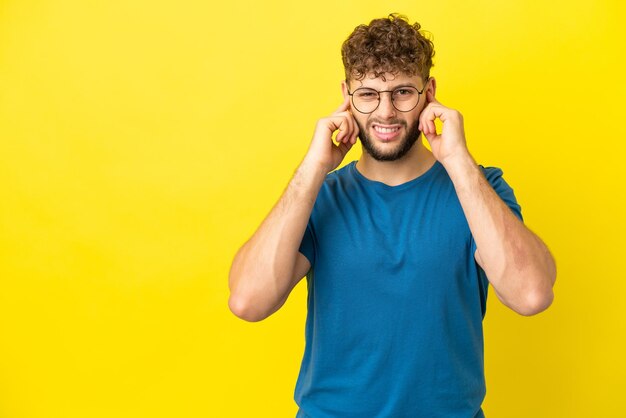 Giovane uomo caucasico bello isolato su sfondo giallo frustrato e che copre le orecchie