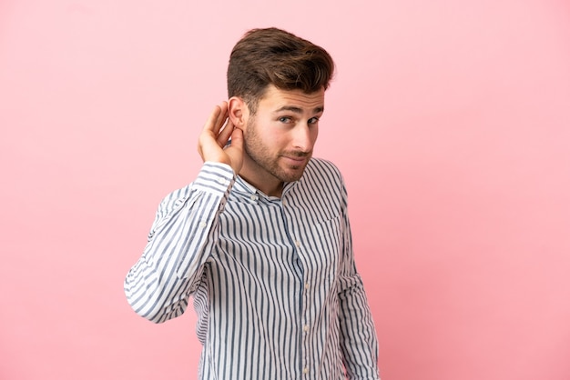 Giovane uomo caucasico bello isolato su sfondo rosa ascoltando qualcosa mettendo la mano sull'orecchio